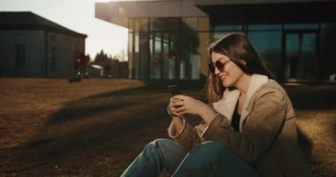 Öğrenci ve teknoloji. Güzel genç bir kadın akıllı telefonuyla bir şeyler okuyor ve modern bir üniversitenin cam binasının önünde güneş gözlüğü takıp gülümsüyor.