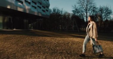 Mutlu genç kız öğrenci akşam güneşinin altında elinde bir çantayla üniversitesine doğru yürüyor. Kampüste.