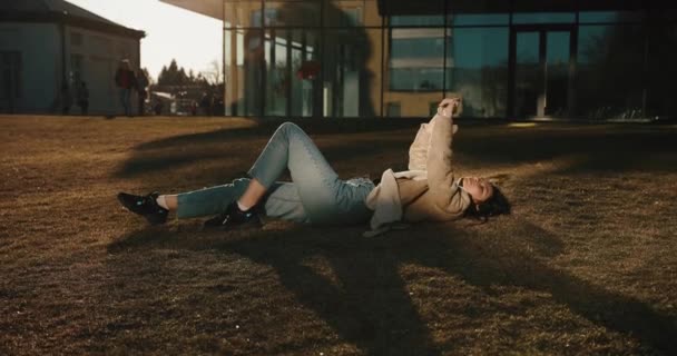 Vibras otoñales. Chica se estira tumbada en el césped verde y disfrutar del sol de la noche antes de la construcción de vidrio moderno de la universidad — Vídeo de stock