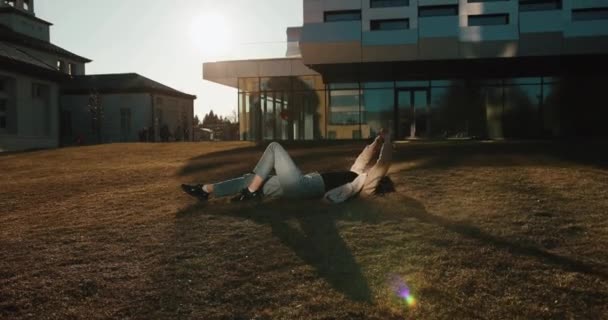 Осенние вибрации. Девушка растягивается на зеленой лужайке и наслаждается вечерним солнцем перед современным стеклянным зданием университета — стоковое видео