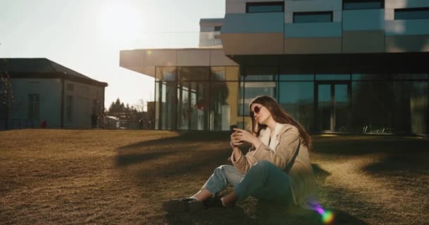 Öğrenci ve teknoloji. Güzel genç bayan, modern bir üniversitenin cam binasından önce güneş gözlüğüyle oturarak akıllı telefonunda bir şeyler okuyor. — Stok video