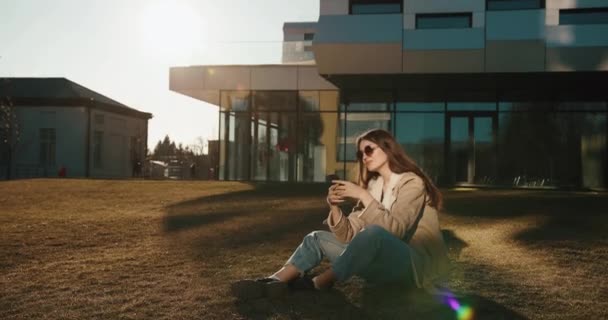 Öğrenci ve teknoloji. Güzel genç bayan, modern bir üniversitenin cam binasından önce güneş gözlüğüyle oturarak akıllı telefonunda bir şeyler okuyor. — Stok video