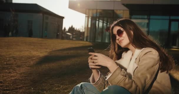 Studenten en technologieën. Mooie jonge vrouw leest iets in haar smartphone zittend in de zonnebril voor het glazen gebouw van een moderne universiteit — Stockvideo