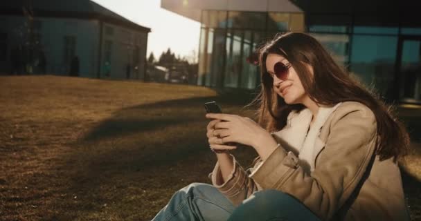 Studenten en technologieën. Mooie jonge vrouw leest iets in haar smartphone en glimlacht teder zittend in de zonnebril voor het glazen gebouw van een moderne universiteit — Stockvideo