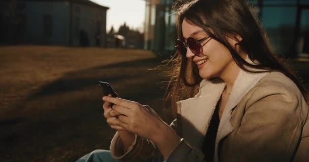 Estudante e tecnologias. Mulher muito jovem digita algo em seu smartphone e sorri concurso sentado nos óculos de sol antes do edifício de vidro de uma universidade moderna — Vídeo de Stock