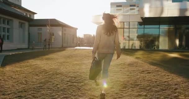 Mira desde atrás a la joven estudiante saltando caminando en los rayos del atardecer hacia el moderno edificio de cristal — Vídeo de stock