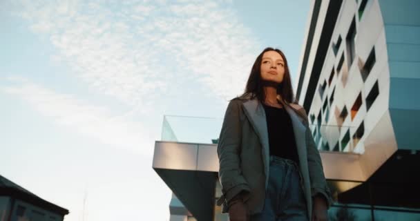 Look from below at confident and serious young woman standing before the modern glass building — Stock Video