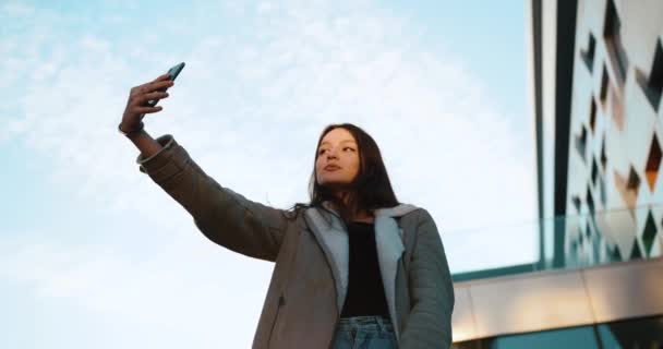Mooie jonge vrouw in moderne casual kleding neemt een selfie staande voor een glazen gebouw in de stralen van de avondzon — Stockvideo