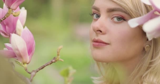 Primer plano de una atractiva joven rubia con hermosos ojos entre flores de magnolia. — Vídeos de Stock