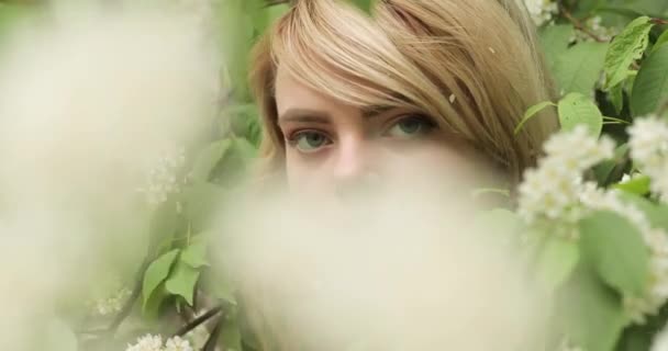 Primer plano de la cara de una joven rubia atractiva entre las ramas de un árbol floreciente. — Vídeos de Stock