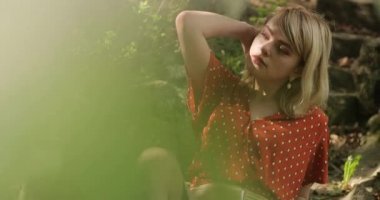 Kırmızı elbiseli çekici bir model yeşil çalıların ve ağaçların arasında poz veriyor.