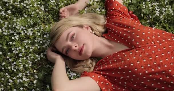 Mladá dívka s blonďatými vlasy, v červených šatech, leží mezi modrými květy zapomenutého léta. věk 20-25 let. — Stock video