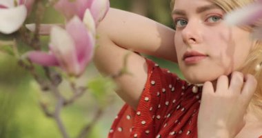 Yakın plan bir portre, doğal sarı saçlı ve mavi gözlü çekici bir kız. Arka planda çiçek açan pembe bir manolya bahçesi. Bahçenin bahar tazeliği.