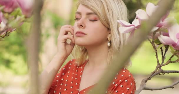 Outdoor close-up portret van jong aantrekkelijk gelukkig glimlachen modieus blond meisje, poseren onder de lente bloeiende magnolia boom. — Stockvideo