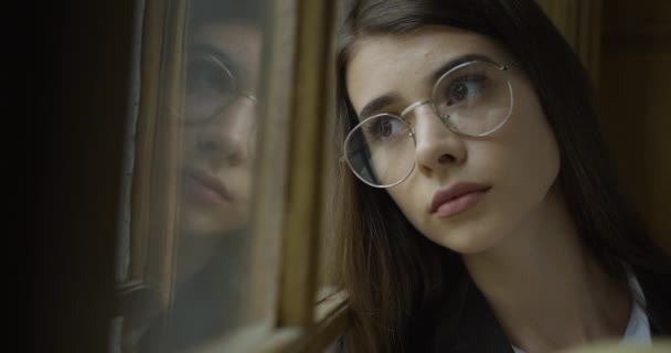 Triste joven en gafas se sienta en el alféizar de la ventana y mira lejos — Vídeos de Stock