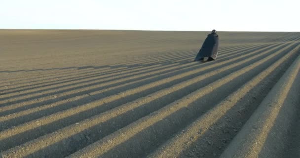 Protección del medio ambiente. Muerte de la naturaleza. Apocalypse. Hombre vestido con capa negra camina a lo largo del campo gris hacia el sol. Vista aérea. — Vídeo de stock