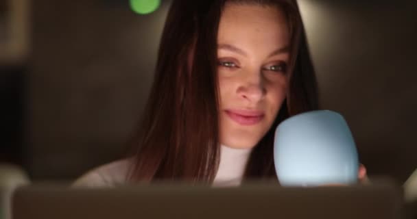 Молодая привлекательная женщина сидит за компьютером и пьет из синей чашки. Работа удаленно из дома — стоковое видео