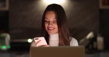 Çekici, serbest çalışan genç bir kadın bilgisayara akıllı telefon kullanıyor ve monitöre bakıyor. Dizüstü bilgisayarda çalışan kadın portresi. Uzaktan kumandalı serbest çalışan evde çalışıyor..