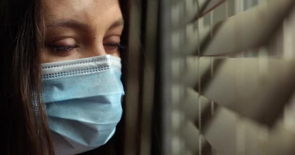 Yeux étroits et visage d'une jeune femme caucasienne portant un masque médical, bougeant les stores avec son doigt. Restez en quarantaine à la maison. — Video
