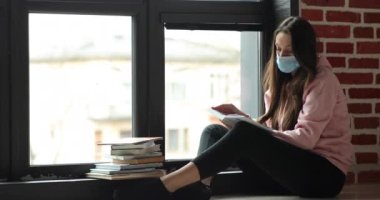 Yüzünde tıp maskesi olan genç bir kız öğrenci pencerenin yanında bir kitap okuyor.