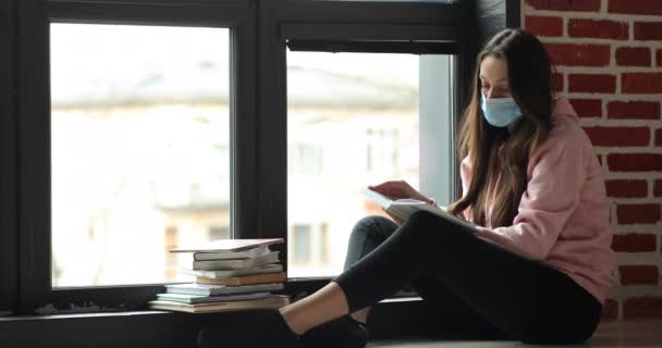 En ung kvinnlig student med mask i ansiktet läser en bok hemma nära fönstret. — Stockvideo