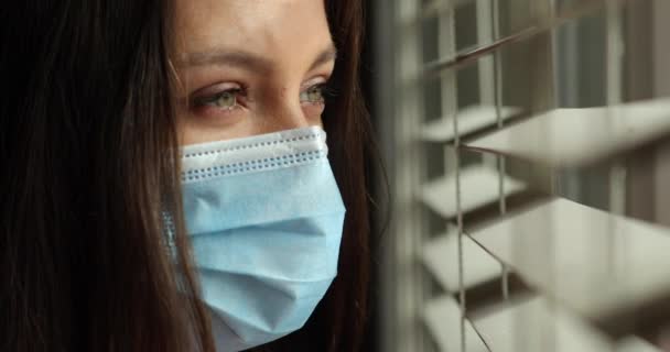 Gros plan portrait d'une belle femme dans un masque médical de protection debout près de la fenêtre et regardant à travers les stores. Tir 4k. Gros plan sur les yeux — Video