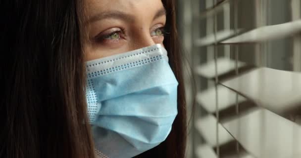 Γυναίκα με μάσκα προσώπου που κοιτάει έξω από το παράθυρο. Νεαρή άρρωστη γυναίκα φαίνεται λυπημένη με προστατευτική μάσκα στο σπίτι πίσω από το παράθυρο σε καραντίνα και κλείδωμα κατά τη διάρκεια Covid — Αρχείο Βίντεο
