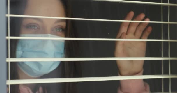 Tıbbi maskeli genç bir kadın pencerenin perdelerinden dışarı bakıyor.. — Stok video