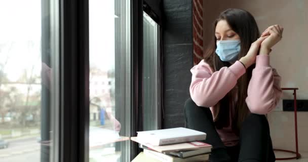 Vista lateral de una atractiva mujer caucásica en una máscara médica sentada en el alféizar de la ventana leyendo un libro y mirando por la ventana 4 k — Vídeo de stock