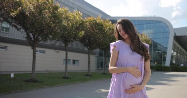 Una giovane donna incinta attraente cammina lungo un vicolo pieno di alberi, guarda nella fotocamera e incanta con il suo aspetto incredibile. Splendida giovane donna con bellissimi occhi verdi e una lunga bruna — Video Stock