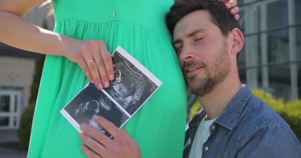 魅力的な若い髭の男は彼の妊娠中の妻の腹を抱擁し、超音波検査の写真を保持しています — ストック動画