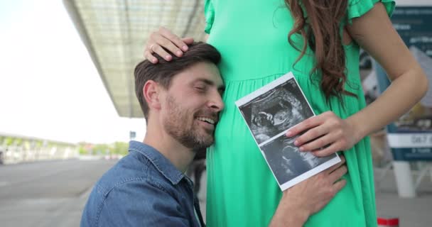 Feliz marido abraza a las esposas embarazadas vientre. Foto de examen por ultrasonido de mujeres embarazadas. — Vídeo de stock