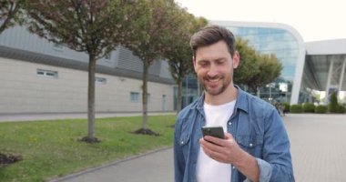 Çekici, genç, sakallı bir adam elinde telefonla caddede yürüyor ve gülümsüyor.