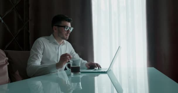 Hombre de negocios con ropa casual trabajando en una computadora portátil y bebiendo café o té — Vídeos de Stock