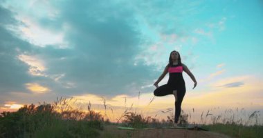 Dışarıda dinlenen genç bir kadın, ağaç pozisyonu alıyor. Bir bacağında balans olan bir kadın. Doğanın ahengi içinde. Ağaç duruşu. Gün batımında yoga yapan kadın..