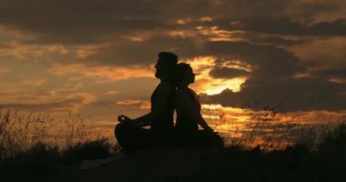 Erkek ve kadın arka arkaya lotus pozisyonunda oturuyorlar. Çift dışarıda yoga yapıyor. Ebedi uyum arayışı. Zihin ve beden. Arka planda güzel doğa, bulutlu gökyüzü ve harika gün batımı..