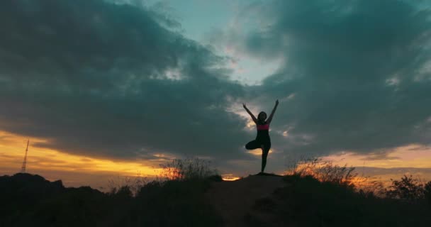 Silhuett av sportig kvinna som står hos yogaposer utomhus. Yogi utövar yogarörelser och positioner i naturen. Idrottare balanserar på ett ben. Vacker himmel och sol som bakgrund. Friska aktiva — Stockvideo