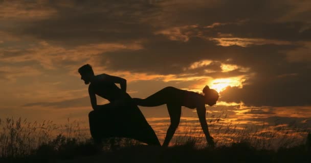 Silhouette eines Mannes und einer Frau beim Yoga. Junges Paar praktiziert Yoga bei Sonnenuntergang. Paar beim Sport im Gras. Zeitlupen-Yoga in der goldenen Stunde.