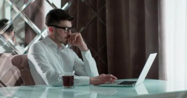 Masada dizüstü bilgisayarla çalışan bir iş adamı. Adam yorgun. Gözlüklerini çıkardı. Masanın üzerinde bir fincan kahve ya da çay var. Arkasında güzel bir ayna duvarı var..