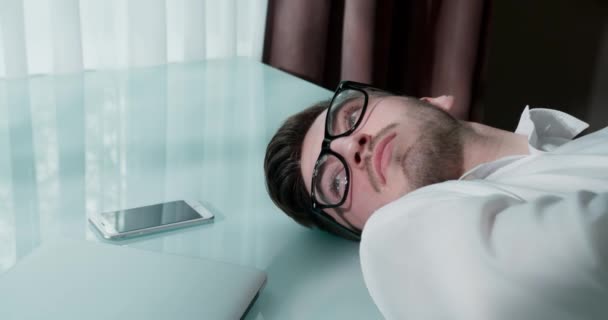 Egy szemüveges férfi fekszik az asztalon laptoppal és okostelefonnal. Becsukja a szemét. Az ember szomorú.. — Stock videók