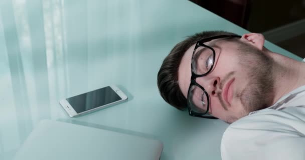 Homem de óculos deitado na mesa com laptop e smartphone. Ele fecha os olhos. — Vídeo de Stock