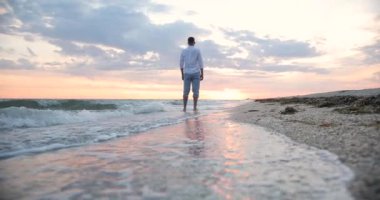 Beyaz gömlekli genç bir adam denizin kenarında duruyor ve güzel gün batımını seyrediyor.