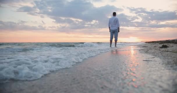Hátul egy fiatal férfi áll a tenger mellett, fehér ingben, és gyönyörű naplementét néz. — Stock videók