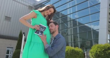 Genç sakallı adam hamile karısına arkadan sarılıp karnını okşuyor.