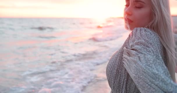 Primer plano de una joven con el pelo largo y rubio de pie junto al mar y disfrutando de los últimos rayos al atardecer. Emoción. — Vídeos de Stock