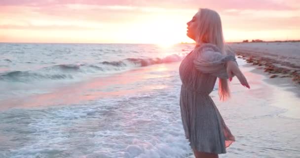 Attraktives stylisches Modell erfreut friedlich das goldene Sonnenlicht über dem Meer. Attraktive Reisende betrachten den Sonnenuntergang am Strand. Blonde Frau genießt Natur im Freien — Stockvideo