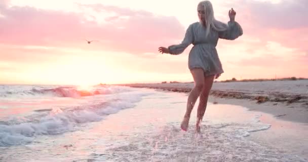 Jong sexy blond Kaukasische vrouw met lang haar in lopen langs de zandige kust bij mooie roze zonsondergang in de zomer in de wind. — Stockvideo
