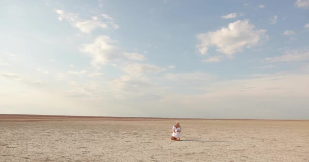 Vonzó fiatal nő szőke hajjal, fehér ruhában, a homokon ülve. A háttérben sok sirály sétál a homokon.. — Stock videók