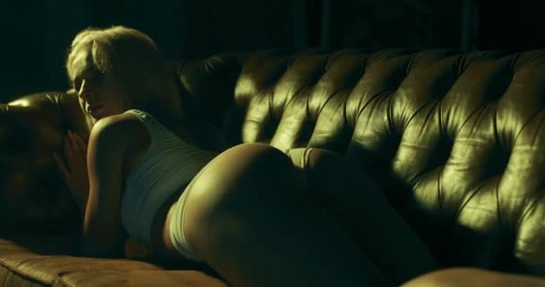 Młoda urocza kobieta uwodzicielsko leży na kanapie w ciemnym pokoju, afiszując się swoim seksownym ciałem. — Wideo stockowe