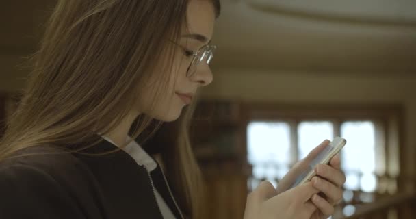 Studente donna, che lavora con lo smartphone. Affascinante ragazza legge qualcosa nel suo telefono in piedi in biblioteca — Video Stock